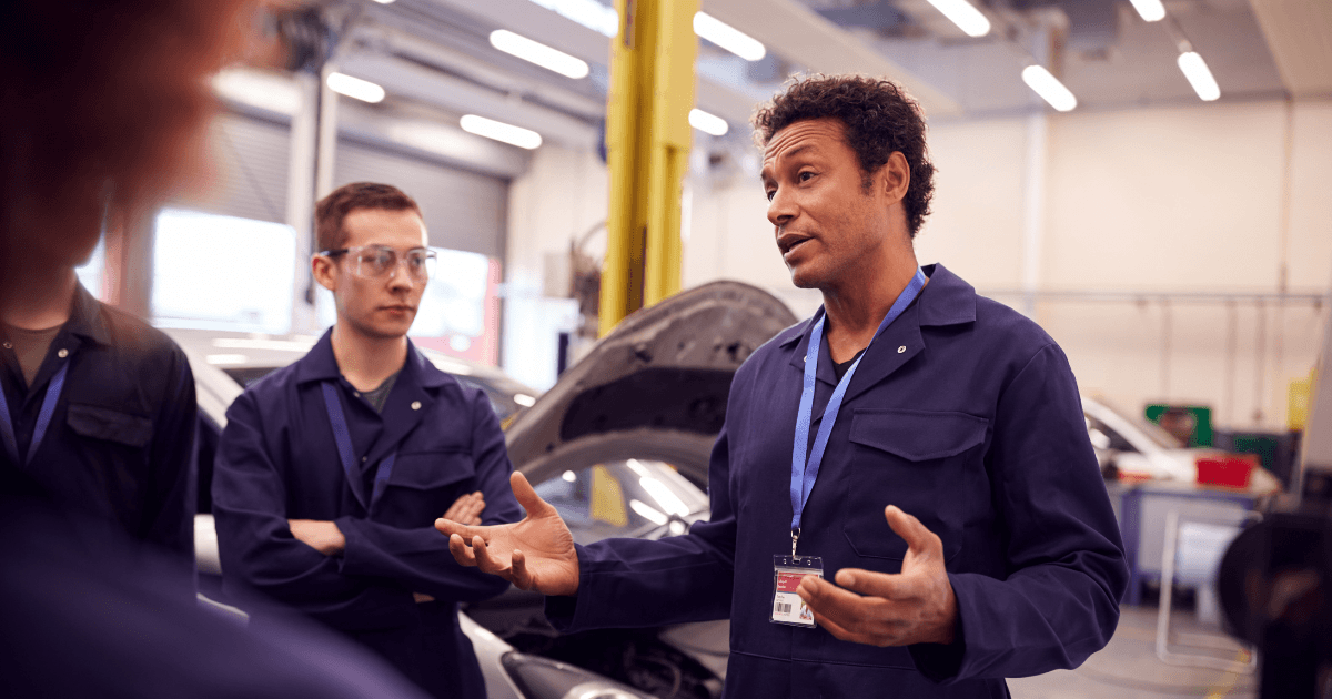 Four workers in an auto shop talk in front of a car being repaired. One older worker appears to be instructing the others. 